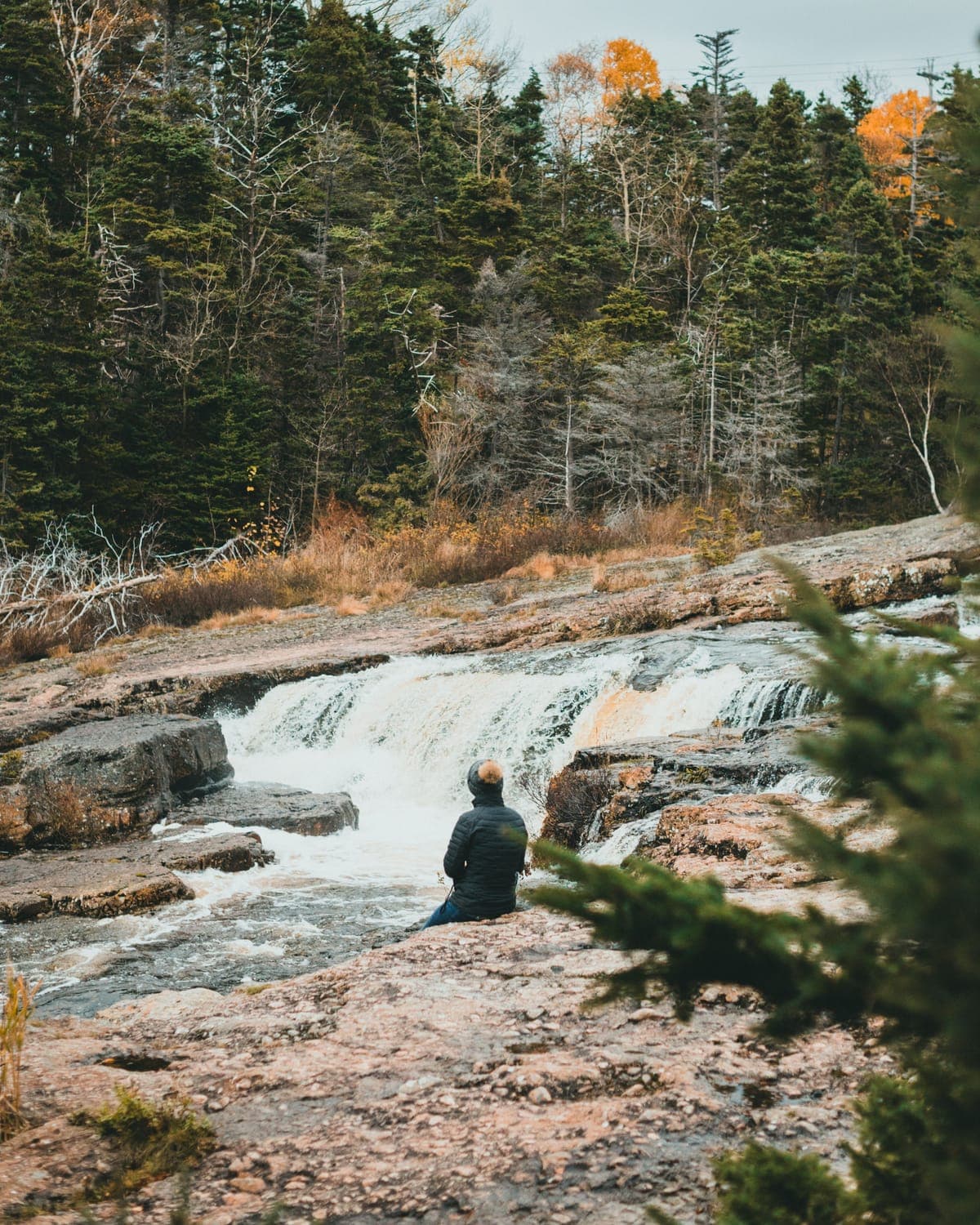 A person sitting alone on flat rock beside a wide, rushing waterfall, autumn woods rising behind
