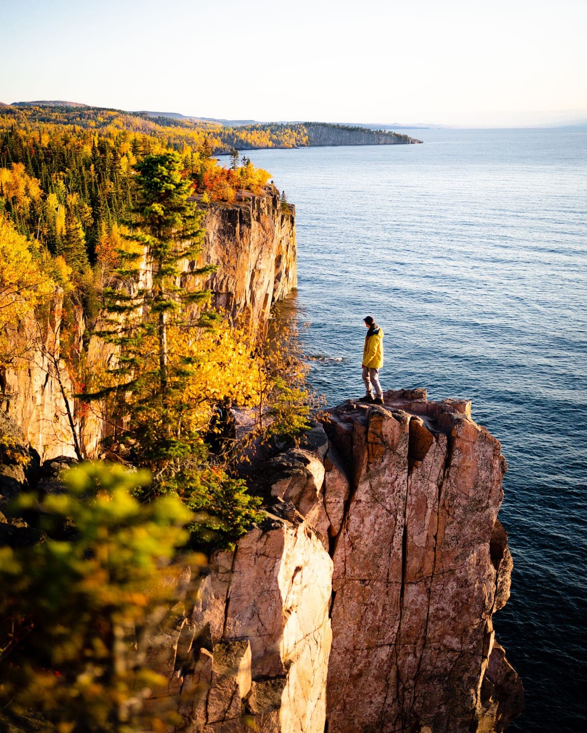 A person standing on a rock outcrop above a wide lake, gold autumn forest along the cliffs behind